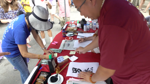 A vendor demonstrates calligraphy at a street fair, while a woman observes and takes notes. Various supplies are on the table.