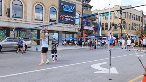 Two players set up a volleyball net on a city street, with a crowd and shops in the background.