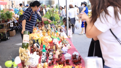 A bustling market scene with vendors selling colorful figurines and plants, and shoppers browsing the stalls.