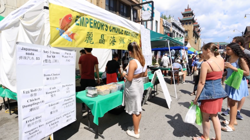 A bustling street market with food stalls, people browsing, and a sign for "Emperor's Choice Restaurant" in Chinatown.