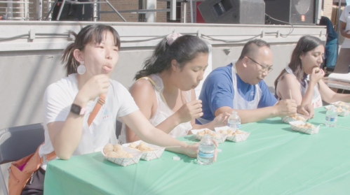 Four people sitting at a table eating from takeout containers, with water bottles in front of them at an outdoor event.