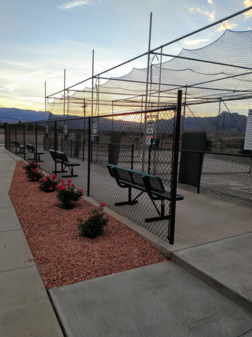 A batting cage area with benches, surrounded by a fence and flower beds, under a sunset sky.