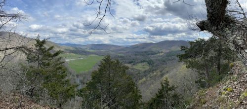 A panoramic view of rolling hills and valleys under a cloudy sky, framed by trees in the foreground.