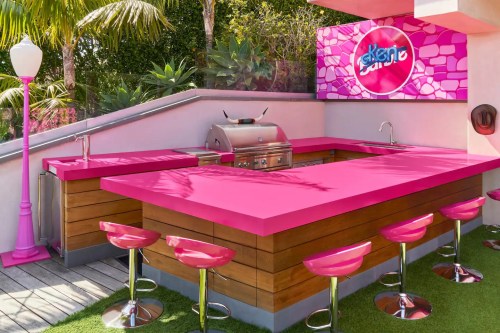 A vibrant outdoor kitchen with a pink countertop, grill, and bar stools, surrounded by greenery.