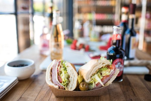 A close-up of a sandwich with layers of meat, lettuce, and tomato, served in a basket with olives, on a wooden table.