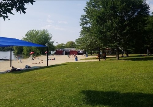 A sandy beach area with people relaxing, trees, and red changing huts near a calm lake under a blue sky.
