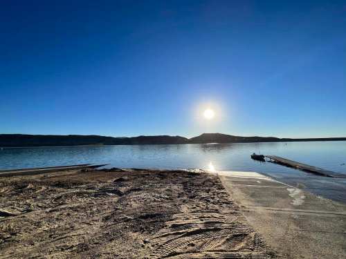 A serene lake at sunrise, with a clear sky and distant mountains reflecting on the water's surface.