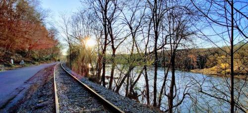 A serene riverside scene with a railway track, bare trees, and a sunset reflecting on the water.