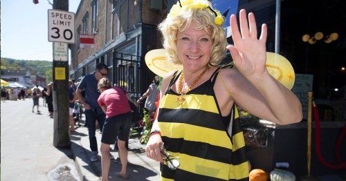 A smiling woman in a bee costume waves at the camera during a lively street festival.
