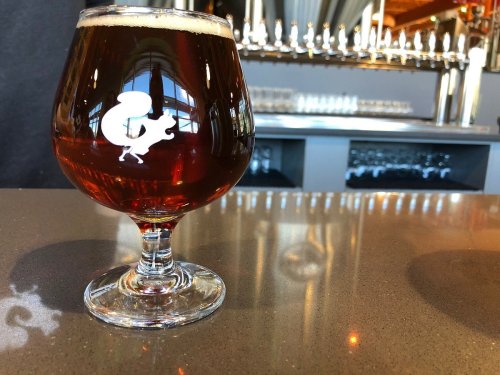 A glass of amber beer sits on a bar counter, reflecting light, with a brewery tap in the background.