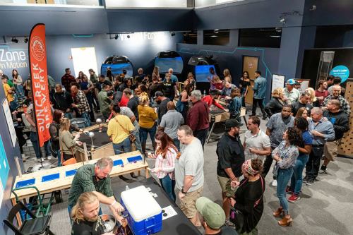 A large crowd gathers in a modern space, enjoying an event with tables, food, and banners in a lively atmosphere.