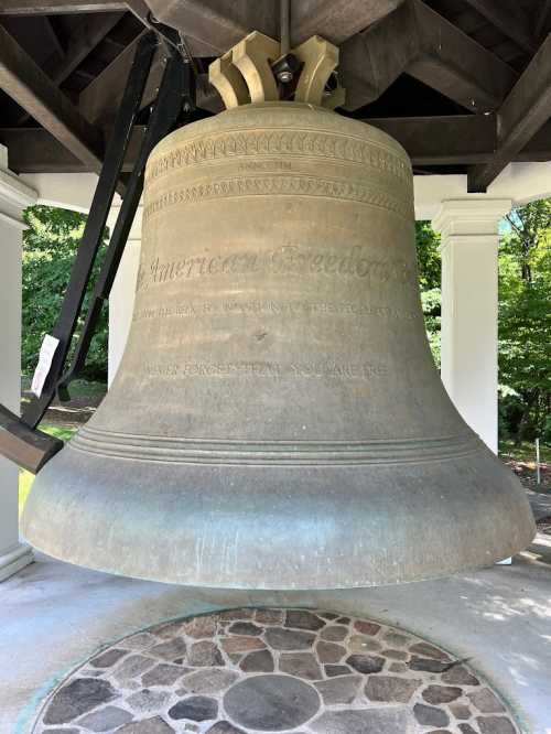 A large bronze bell hangs in a pavilion, inscribed with "America's Freedom." Surrounded by greenery.