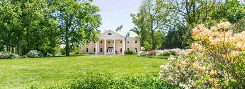 A large, elegant yellow mansion surrounded by lush green grass and blooming flowers on a sunny day.