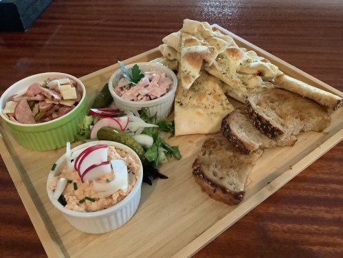 A wooden platter with dips, sliced bread, crackers, and a side salad, featuring various toppings and garnishes.