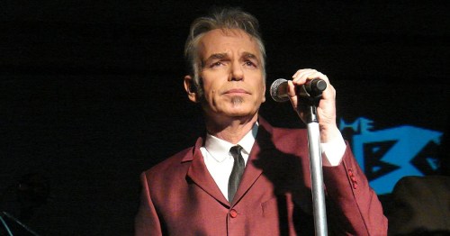 A man in a maroon suit holds a microphone, performing on stage with a focused expression.