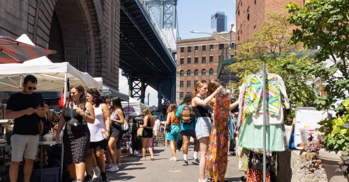 A bustling outdoor market under a bridge, with people browsing stalls and colorful clothing on display.