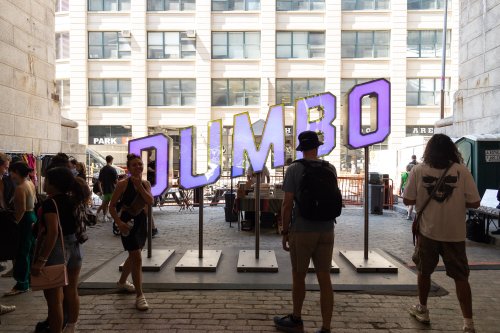 A vibrant "DUMBO" sign stands in an urban space, with people browsing nearby stalls and shops.