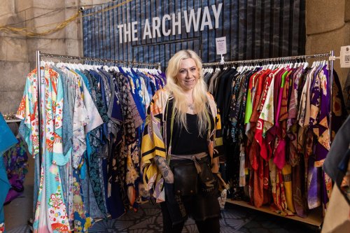 A smiling person stands in front of a rack of colorful clothing at a market, with "THE ARCHWAY" sign in the background.