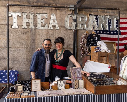 A man and woman stand behind a market stall displaying jewelry and accessories, with a sign reading "THEA GRANT."