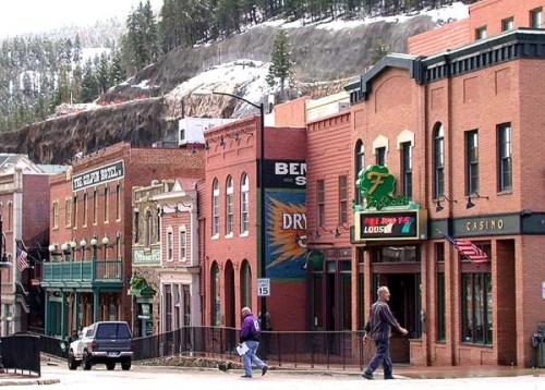 Historic buildings line a street in a small town, with snow-capped hills in the background and people walking by.