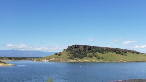 A serene landscape featuring a calm lake, green hills, and a rocky outcrop under a clear blue sky.