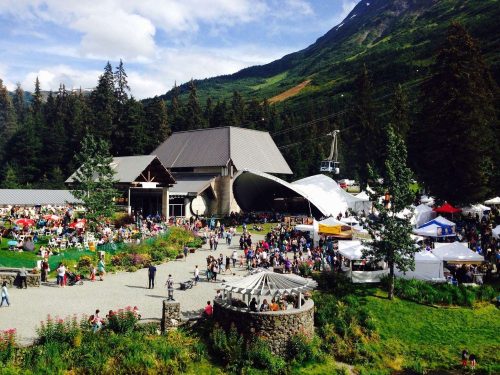 A bustling outdoor festival with crowds, tents, and a scenic mountain backdrop. Trees and buildings are visible.