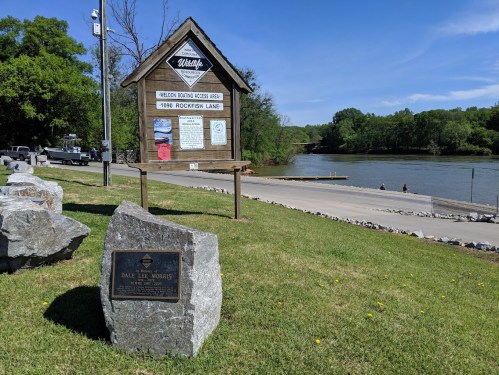 A scenic view of Rockfish Lake with a wooden sign and a plaque in the foreground, surrounded by greenery.