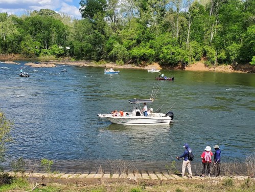 A river scene with several boats and people fishing along the shore, surrounded by lush green trees.