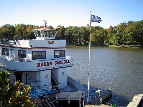 A boat named "Madam Carroll" docked by a calm lake, with trees in autumn colors and a pirate flag flying.