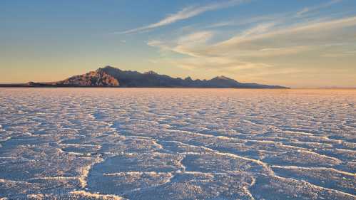 A vast salt flat stretches towards distant mountains under a clear sky, with textured patterns on the ground.