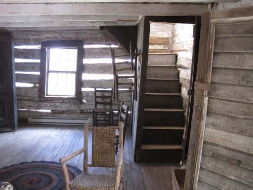 Interior of a rustic log cabin featuring wooden stairs, a woven chair, and a round rug on the floor.
