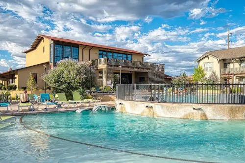 A sunny pool area with lounge chairs, surrounded by greenery and a modern building under a blue sky with clouds.