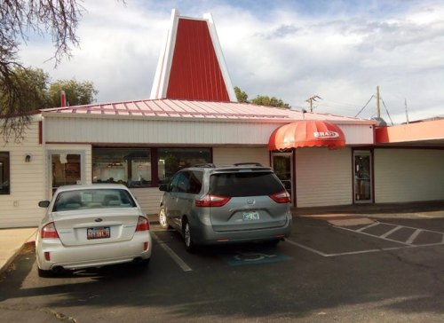 A fast-food restaurant with a red roof and awning, featuring parked cars in front and a cloudy sky above.