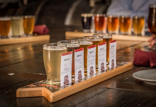 A wooden flight board with six small glasses of beer, each labeled, on a rustic table with more drinks in the background.