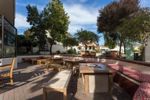 Outdoor seating area with wooden benches, a fire pit, and lush greenery under a blue sky.