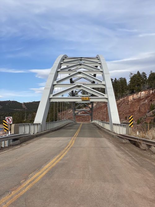 A modern arch bridge spans a road, surrounded by trees and hills under a partly cloudy sky.
