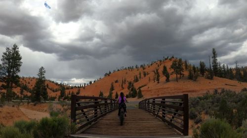 A person on a bike crosses a wooden bridge, surrounded by orange hills and dark, cloudy skies.