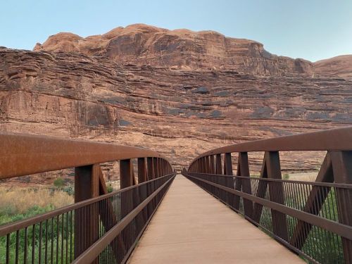 A metal bridge leads through a rocky landscape, flanked by towering red rock formations under a clear sky.