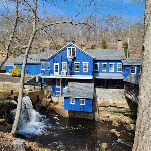A blue building by a stream with a small waterfall, surrounded by trees and a clear blue sky.