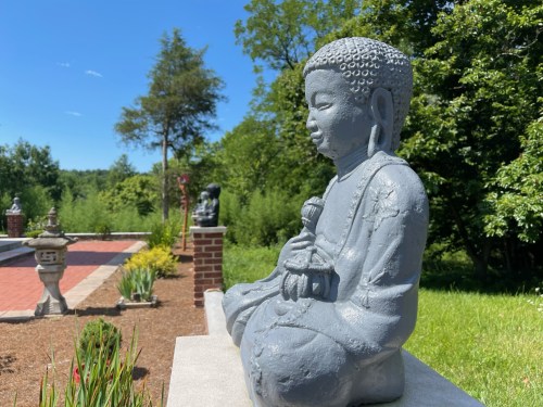 A serene Buddha statue sits in a garden, surrounded by greenery and stone features under a clear blue sky.