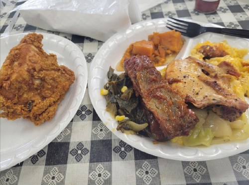 Two plates of food: one with fried chicken, the other with meat, greens, and sweet potatoes on a checkered tablecloth.