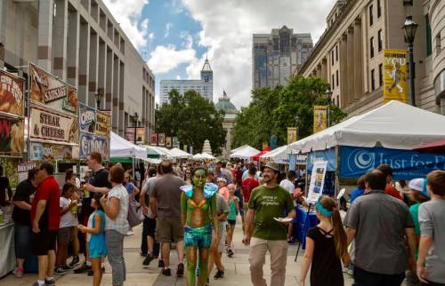 A bustling street fair with food stalls, people enjoying the event, and a performer in body paint among the crowd.