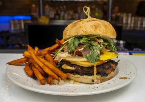 A large burger topped with greens, cheese, and a fried egg, served with a side of sweet potato fries.