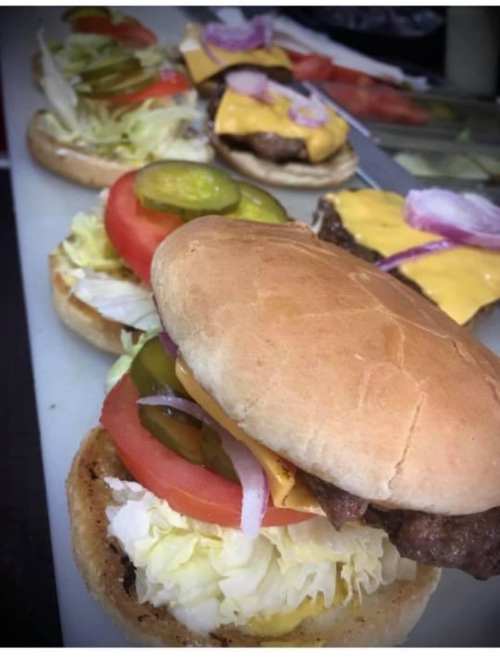 A close-up of a burger with lettuce, tomato, pickles, and cheese, with additional burgers lined up in the background.