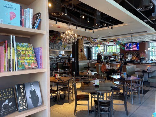 A cozy restaurant interior with wooden tables, bookshelves, and colorful decor, featuring diners and a chandelier.
