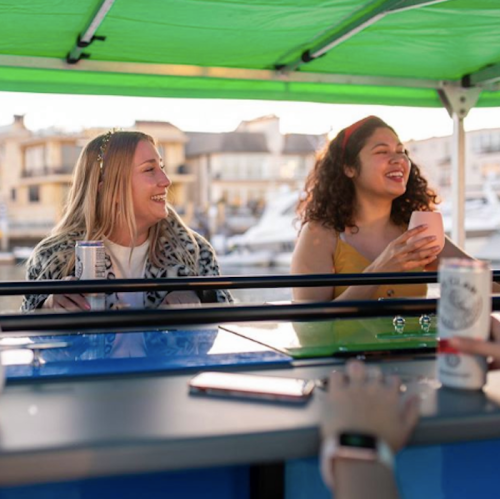 Two smiling women enjoy drinks at a bar under a green canopy, with boats visible in the background.