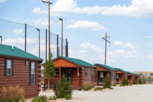 A row of rustic log cabins with green roofs, set against a blue sky with scattered clouds and utility poles in the background.