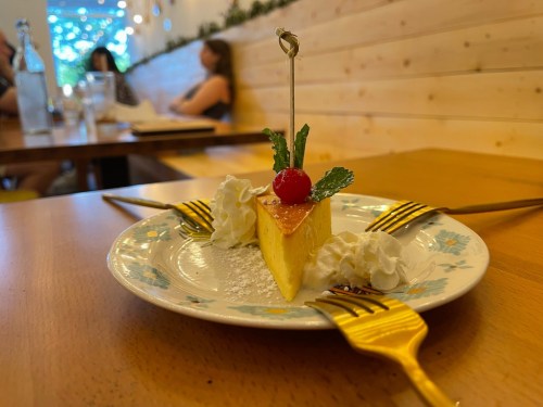 A slice of cheesecake topped with a cherry and mint, served on a decorative plate with three gold forks.