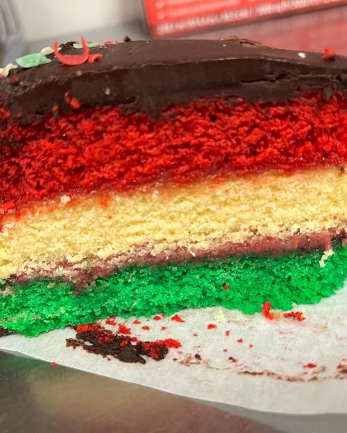 A close-up of a slice of rainbow cake with red, yellow, and green layers, topped with chocolate frosting.