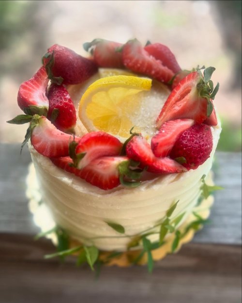 A beautifully decorated cake topped with strawberries and a lemon slice, surrounded by green leaves.